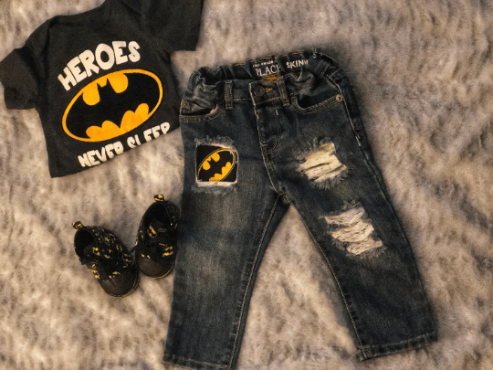 A pair of distressed blue jeans for kids with a Batman logo on one leg, accompanied by a black shirt with the text 'Heroes NEVER SLEEP' and a pair of baby shoes.