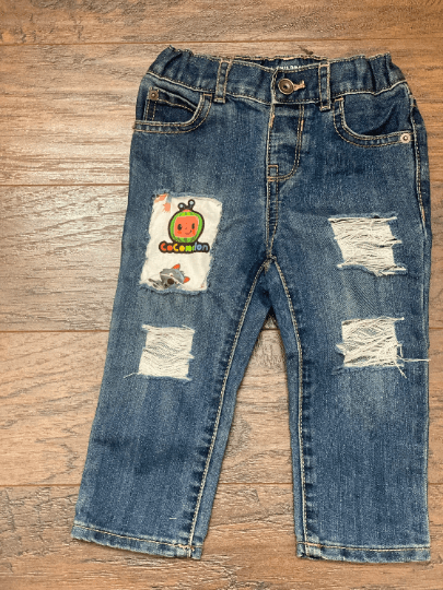 A pair of distressed blue denim skinny jeans with a Cocomelon cartoon character sticker on one leg, displayed on a wooden floor.