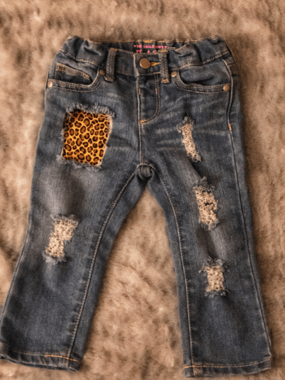 A pair of distressed denim jeans for kids with leopard print on one leg and various other designs such as a cheetah print on the other, placed on a soft surface.