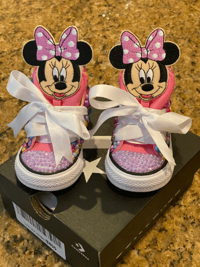 Custom pink Converse sneakers with Minnie Mouse design, featuring bling rhinestones and pearls, displayed on a black platform with a bow and ribbon laces.